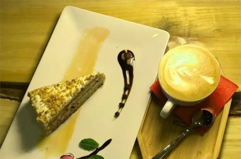 یک برش کیک خوش‌طعم با تزئین مغز، در کنار فنجان لاته آرت زیبا و دلنشین، نشان‌دهنده کیفیت منوی کافی شاپ اچ اند اچ برای لحظاتی شیرین.