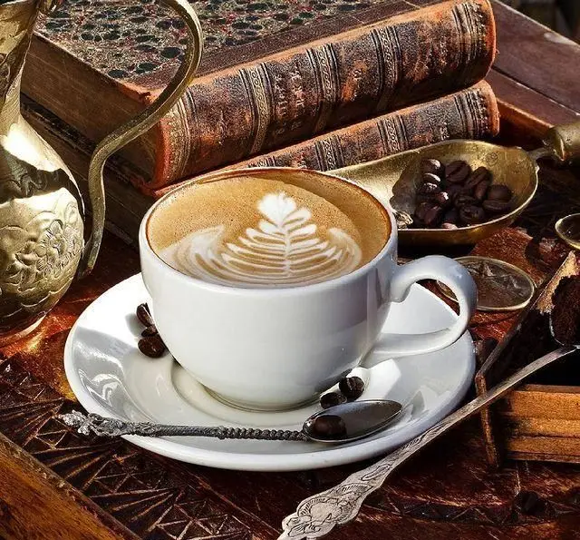 یک فنجان قهوه لاته آرت زیبا، در کنار کتاب‌های قدیمی، قوری برنجی، دانه قهوه، قاشق‌های آنتیک و دسر در کافه خلبان.