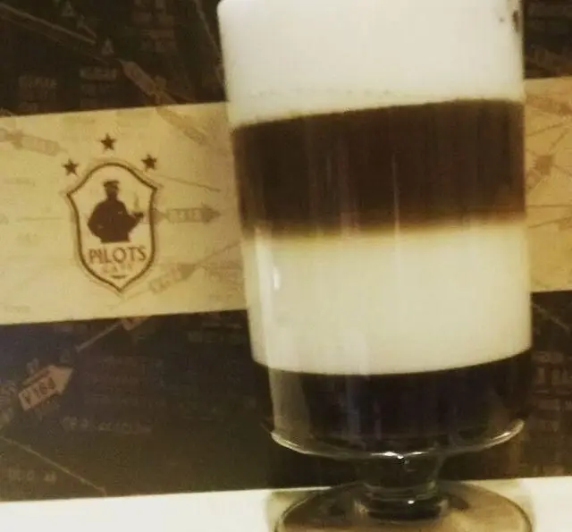 قهوه لایه ای کافه خلبان، نوشیدنی خاص و محبوب با لایه‌های جذاب فوم، شیر و قهوه، در فضایی دلنشین همراه با لوگوی Pilot's Cafe.