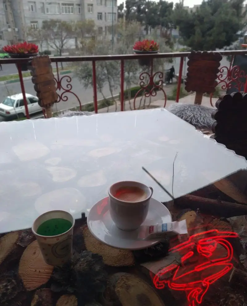 تصویری دلپذیر از بالکن کافه رها، نمایشگر یک فنجان قهوه اسپرسو و نوشیدنی سبز روی میز چوبی با طرح کنده درخت و منظره شهری.