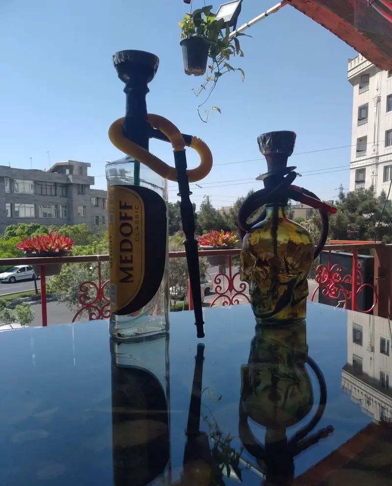 نمایی از فضای باز کافه رها با قلیان‌های خاص (بطری مدوف و طرح اسکلت) روی میز شیشه‌ای. منظره دلنشین شهری، انتخابی عالی برای آرامش.