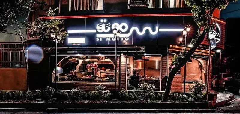 تصویری از نمای بیرونی فست فود سیموتو در شب با تابلوی نئونی "Si Morto" روشن. ورودی اصلی و درختان در اطراف رستوران دیده می‌شوند.