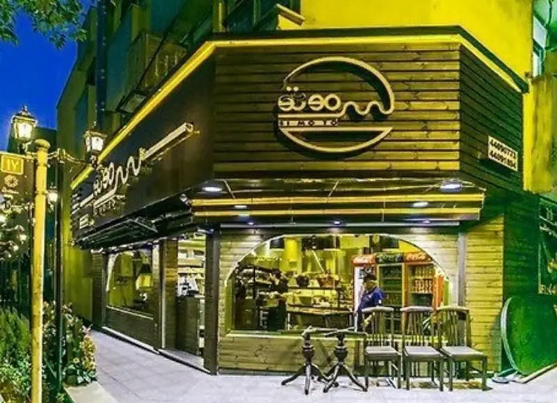 نمای خارجی شب فست فود سیموتو، با طراحی دکوراسیون چوبی مدرن و تابلو نئون لوگوی خلاقانه برگر. مکانی دلنشین برای سرو غذای آماده و لذت از محیط.