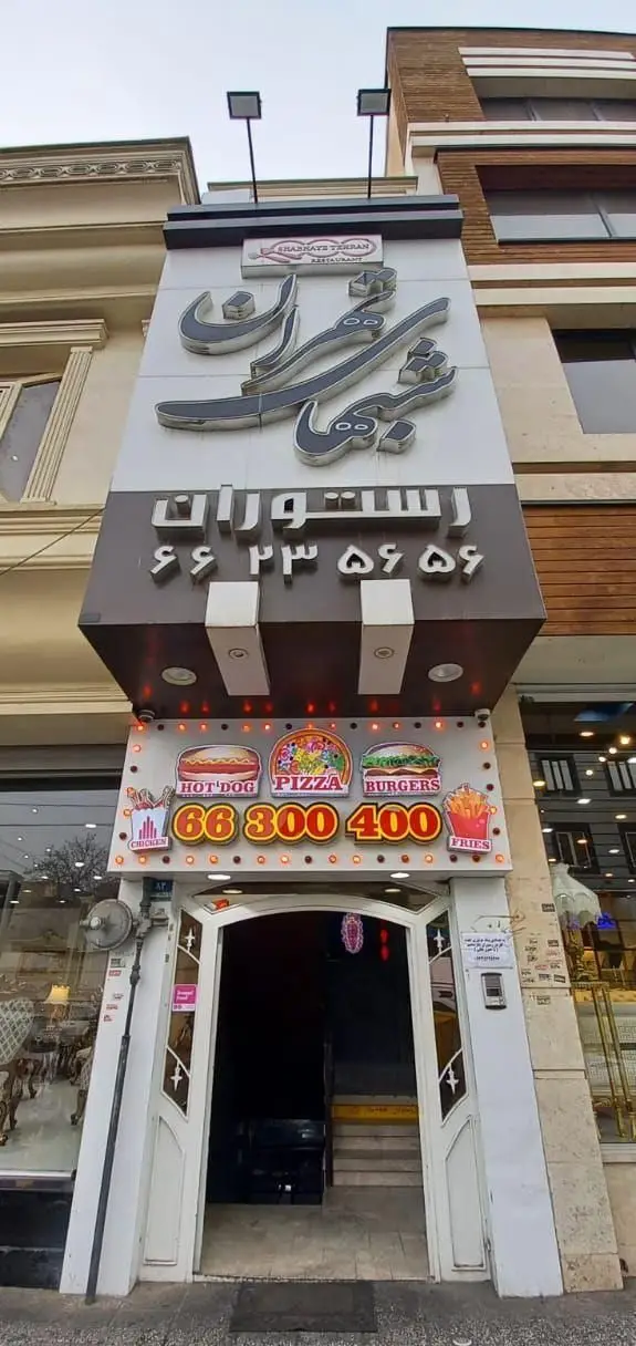 نمای بیرونی رستوران و فست فود شب‌های تهران با تابلوهای درخشان پیتزا، برگر، هات داگ و مرغ سوخاری. شماره‌های تماس: 66235659 و 66300400.
