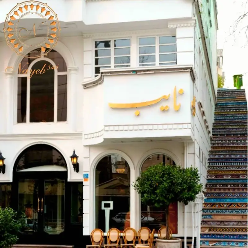 رستوران نایب ساعی با نمای سفید و لوکس، تابلوی طلایی «نایب»، ورودی شیک، میز و صندلی‌های فضای باز و پله‌های کاشی‌کاری شده.