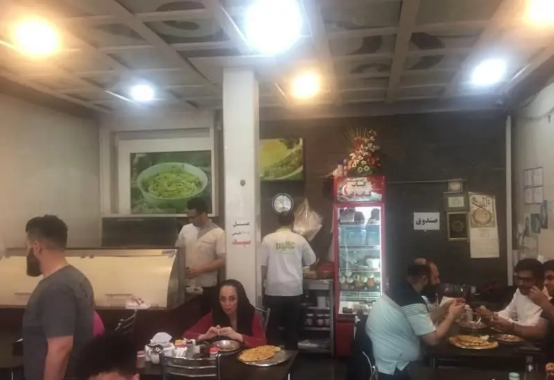 فضای داخلی رستوران ایرانی املت و دیزی سرای علی با میز و صندلی، مشتریان، تصاویر غذاهای ایرانی و کباب، یخچال نوشیدنی و صندوق.