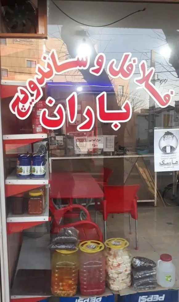 مغازه فست فود باران با تابلو فلافل و ساندویچ، ویترین پر از ترشیجات و فضای داخلی با میز و صندلی قرمز.