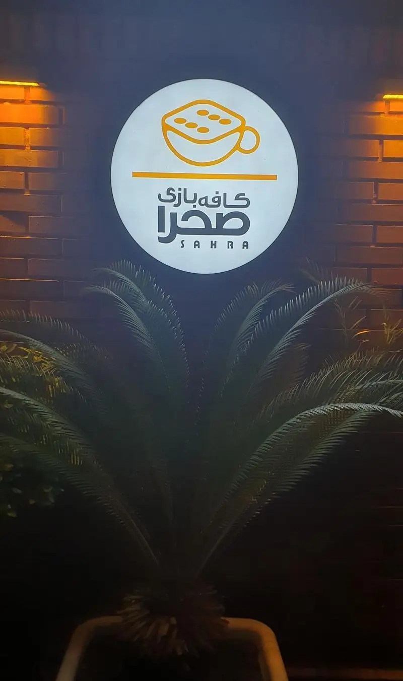 تصویری شبانه از تابلوی گرد و روشن کافه بازی صحرا، با لوگوی فنجان قهوه و تاس، روی دیوار آجری و همراه با گیاه نخل در جلو.