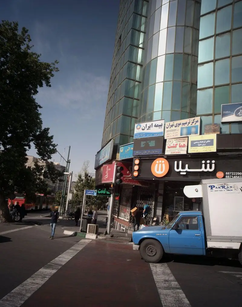 نمای شهری پر جنب و جوش از فست فود شیلا در خیابانی در تهران، شامل تابلوی نارنجی شیلا، ساختمان مدرن، مغازه‌های دیگر و وانت آبی در حال پارک.