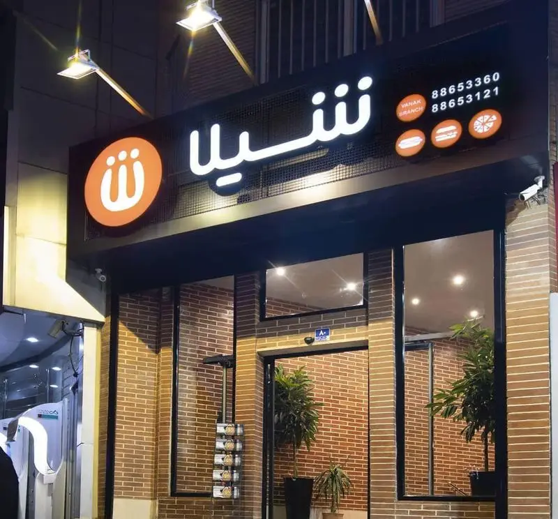 تصویری از نمای شب فست فود شیلا شعبه ونک با تابلو روشن شامل لوگو، تلفن‌های تماس و آیتم‌های منو مثل ساندویچ، برگر و پیتزا.