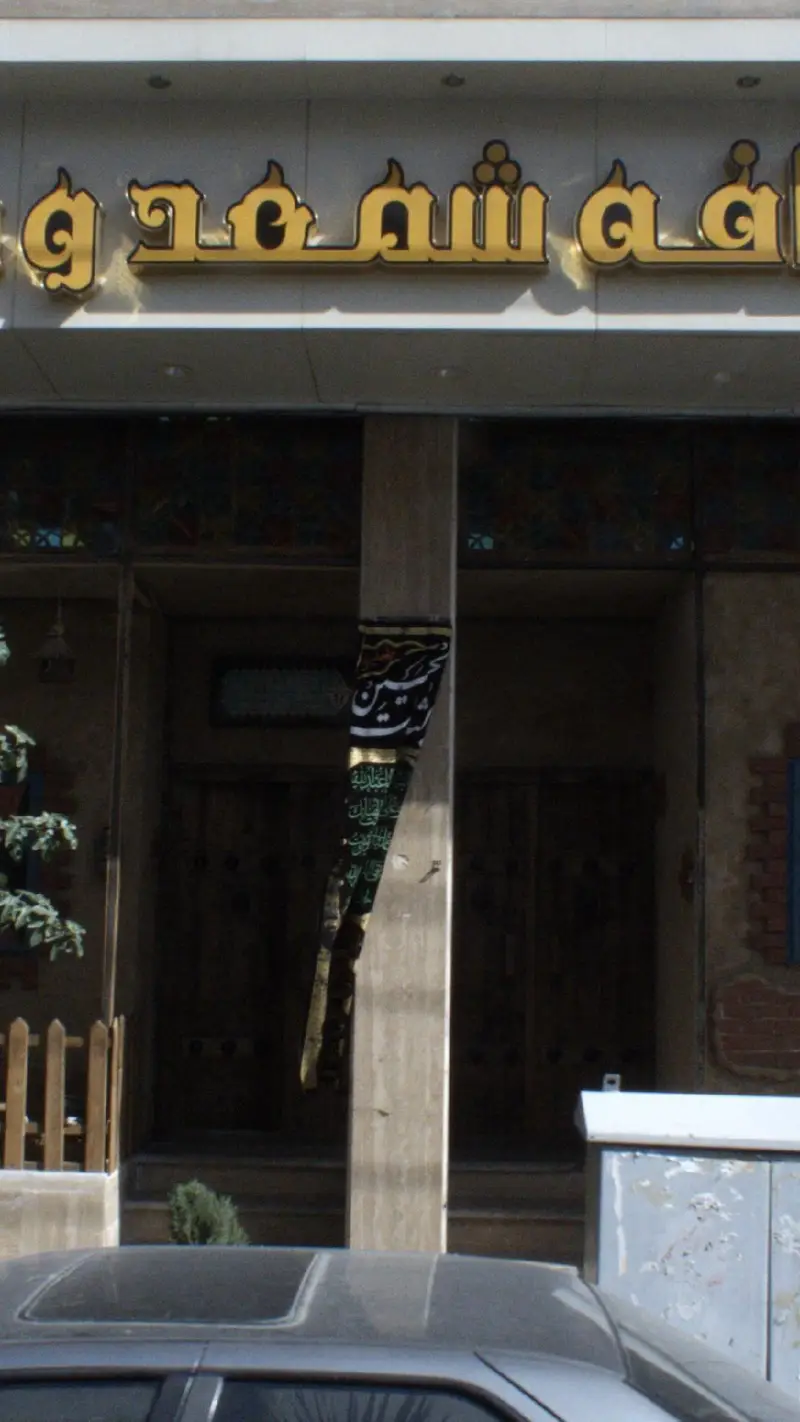 تصویری از نمای خارجی کافه شمعدونی با تابلوی طلایی درخشان و ورودی آن. یک بنر مذهبی نیز روی ستون جلوی کافه نصب شده است.