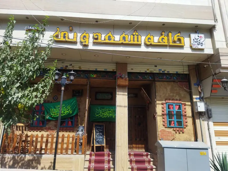 نمای کامل بیرونی کافه شمعدونی با تابلوی بزرگ زردرنگ، پرچم‌های سبز مزین به خطاطی و دکوراسیون جذاب سنتی شامل پنجره‌های آبی و نرده چوبی.