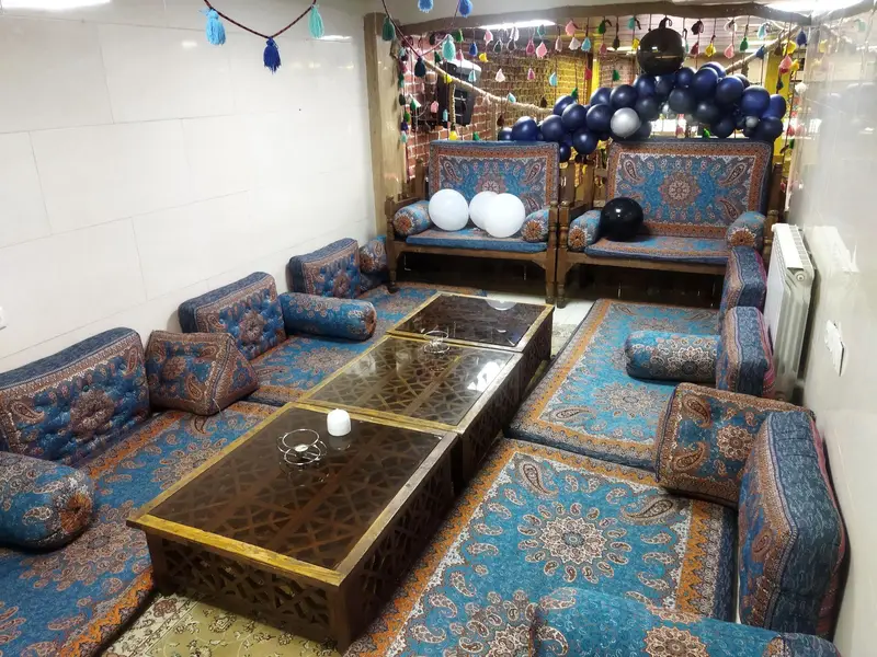 فضای داخلی دنج و سنتی کافه شمعدونی با مبلمان آبی طرح‌دار، پشتی‌ها و تشک‌های زیبا، میزهای چوبی و دکوراسیون بادکنکی رنگی.