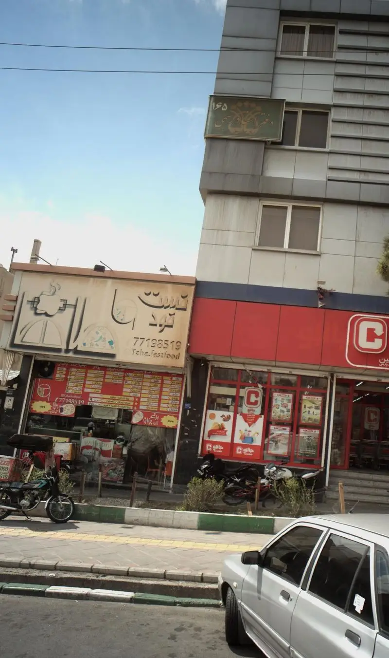 نمای بیرونی رستوران 25 میل (۲۵ میل) در یک روز آفتابی، با تابلوی قرمز رنگ و لوگوی C. فست فود طاها در کنار آن قرار دارد. پیاده‌رو، ماشین و موتور سیکلت پارک شده نیز دیده می‌شود.