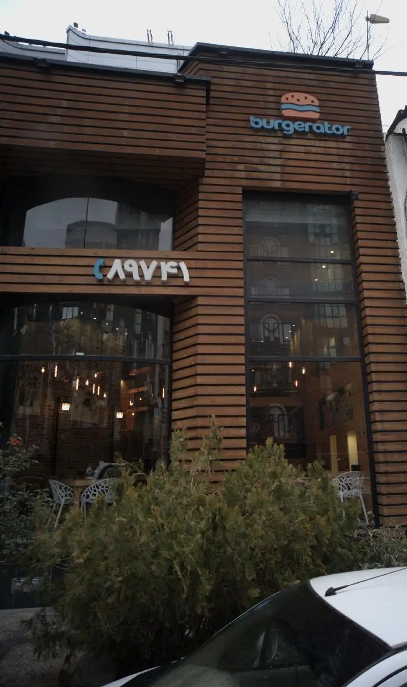 نمای بیرونی فست فود برگراتور با تابلوی آبی رنگ و طرح همبرگر، ساختمان با نمای چوبی و پنجره‌های بزرگ شیشه‌ای.