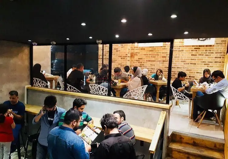 فضای داخلی شلوغ فست فود برگراتور، مشتریان در حال صرف غذا و گفتگو، با دکوراسیون دیوارهای آجری و میزهای چوبی مدرن.