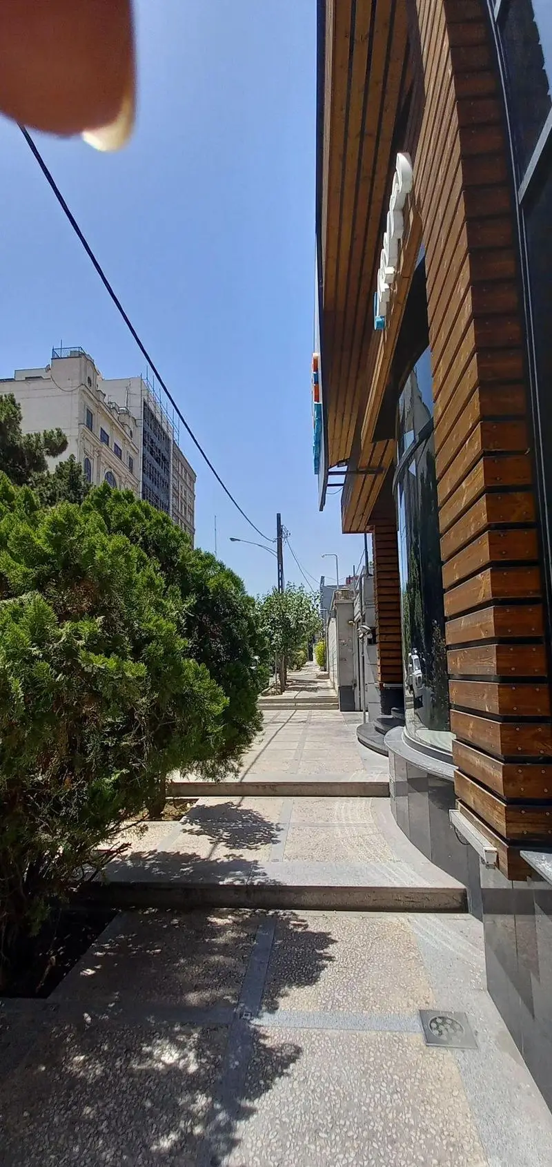 نمای بیرونی فست فود برگراتور با معماری چوبی و شیشه‌ای مدرن. پیاده‌رو، درختان سرسبز و سایه‌های دلنشین در یک روز آفتابی.