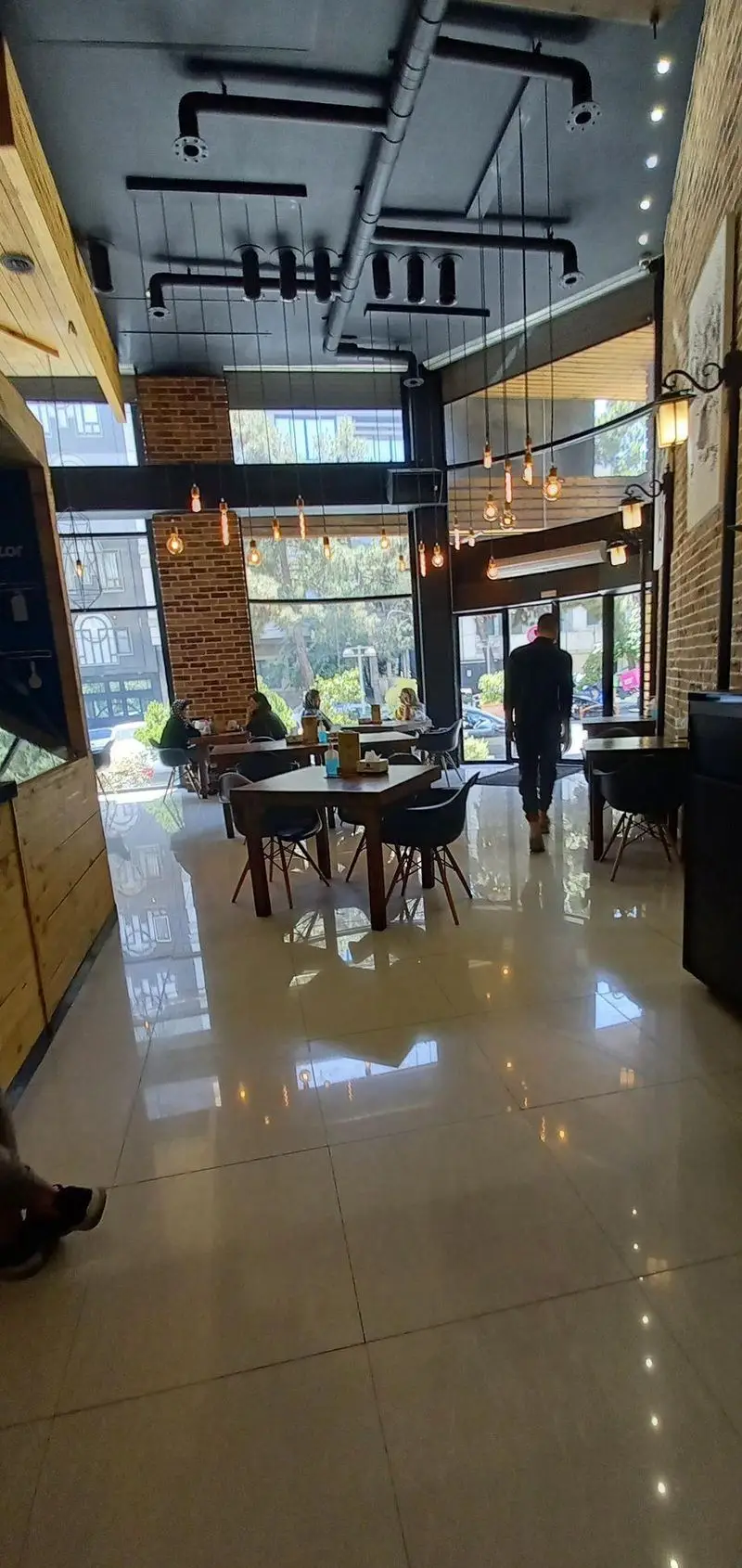 محیط داخلی فست فود برگراتور با طراحی مدرن صنعتی، سقف لوله‌کشی و نورپردازی خاص، میز و صندلی‌های شیک و مشتریان در حال غذا خوردن، بازتاب نور روی کف براق.