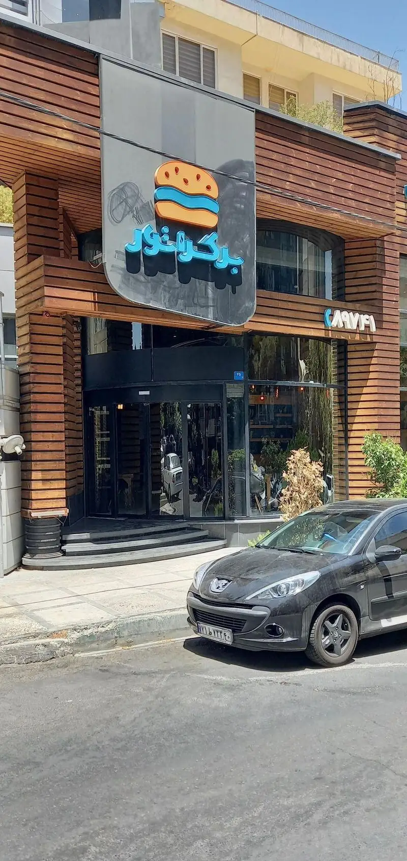 فست فود برگراتور: نمای جلوی ساختمان مدرن با پنل‌های چوبی، ورودی شیشه‌ای بزرگ، و تابلوی درخشان با لوگوی برگر و نام فارسی "برگراتور".