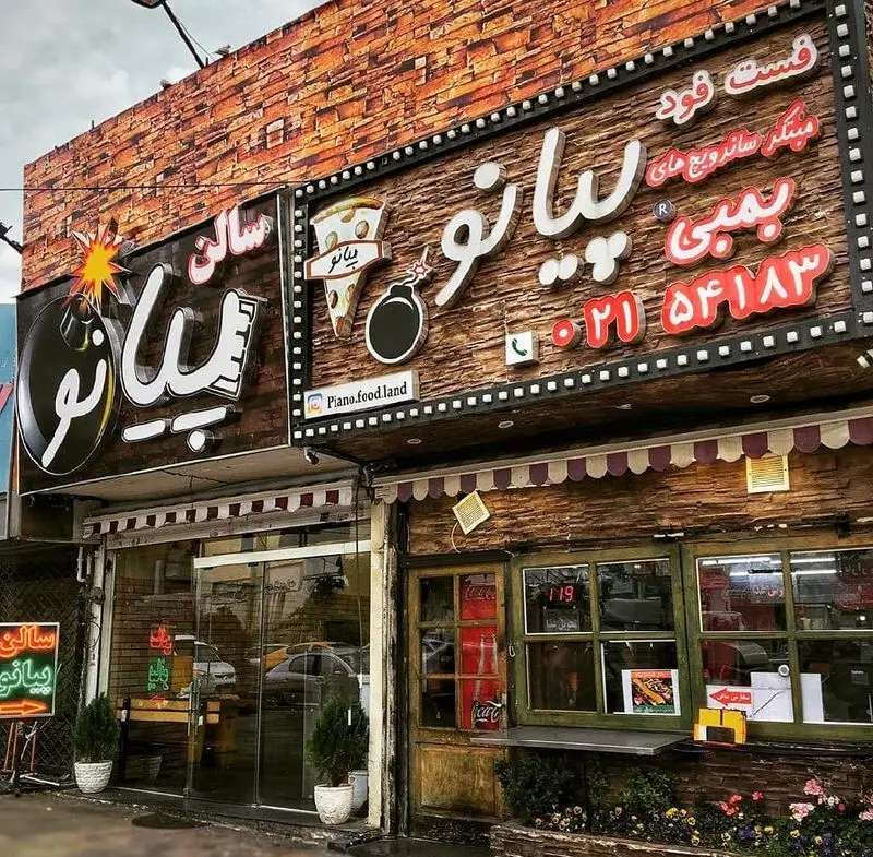 نمای خارجی فست فود پیانو با دو تابلوی بزرگ، لوگوی بمب و پیتزا، ارائه دهنده میکس ساندویچ‌های بمبی و سالن غذاخوری شیک.