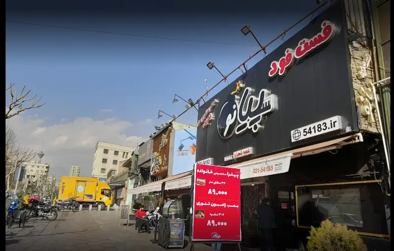 نمای فست فود پیانو در خیابان شلوغی در تهران، با تابلوی درخشان، منوی پیتزا و بمب ژامبون، و اطلاعات تماس ۰۲۱-۵۴۱۸۳.