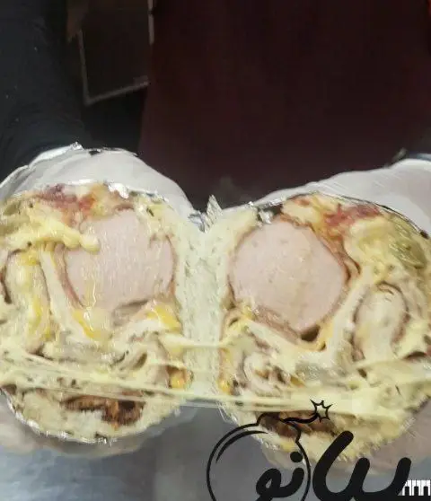 تصویر یک رول ساندویچ هات داگ پنیری و پرملات فست فود پیانو که از وسط برش خورده و پنیر کش‌دار آن دیده می‌شود.