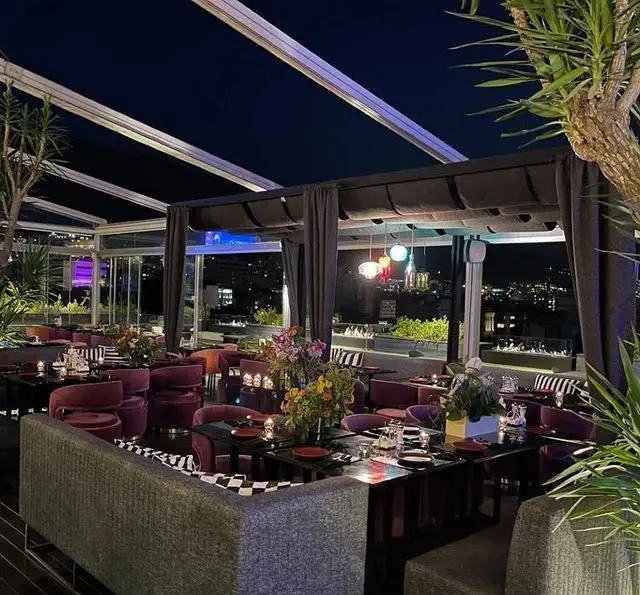 رستوران 10forever: فضای روف گاردن لوکس در شب با سقف شیشه‌ای، چیدمان مبلمان بنفش، و چشم‌انداز خیره‌کننده شهر برای شامی دل‌انگیز.