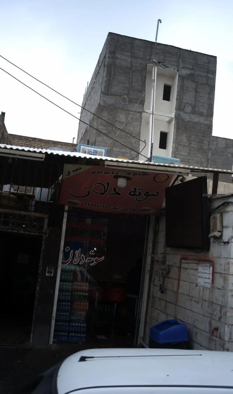 ساندویچی سوته دلان: نمای بیرونی یک مغازه فست فود با تابلوی قرمز و ورودی باز، ساختمان بتنی در پس زمینه و سیم‌های برق.