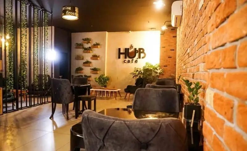 فضای داخلی کافه هاب با طراحی مدرن، دیوار آجری، دکوراسیون گیاهی، مبلمان راحت و لوگوی بزرگ کافه هاب.