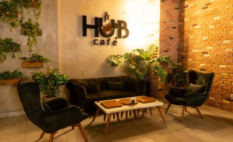 تصویری از محیط دنج و زیبای کافه هاب با دیوارهای آجری و سمنتی، مبلمان راحتی مخملی سبز، گلدان‌های گیاه و میز چوبی قهوه‌خوری.