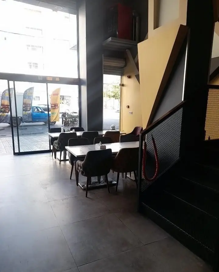 فضای داخلی شیک و مدرن فست فود تیپ تاپ با میز و صندلی، نور طبیعی فراوان و نمایی به بیرون از طریق پنجره‌های بزرگ. مناسب برای سفارش و صرف غذا.