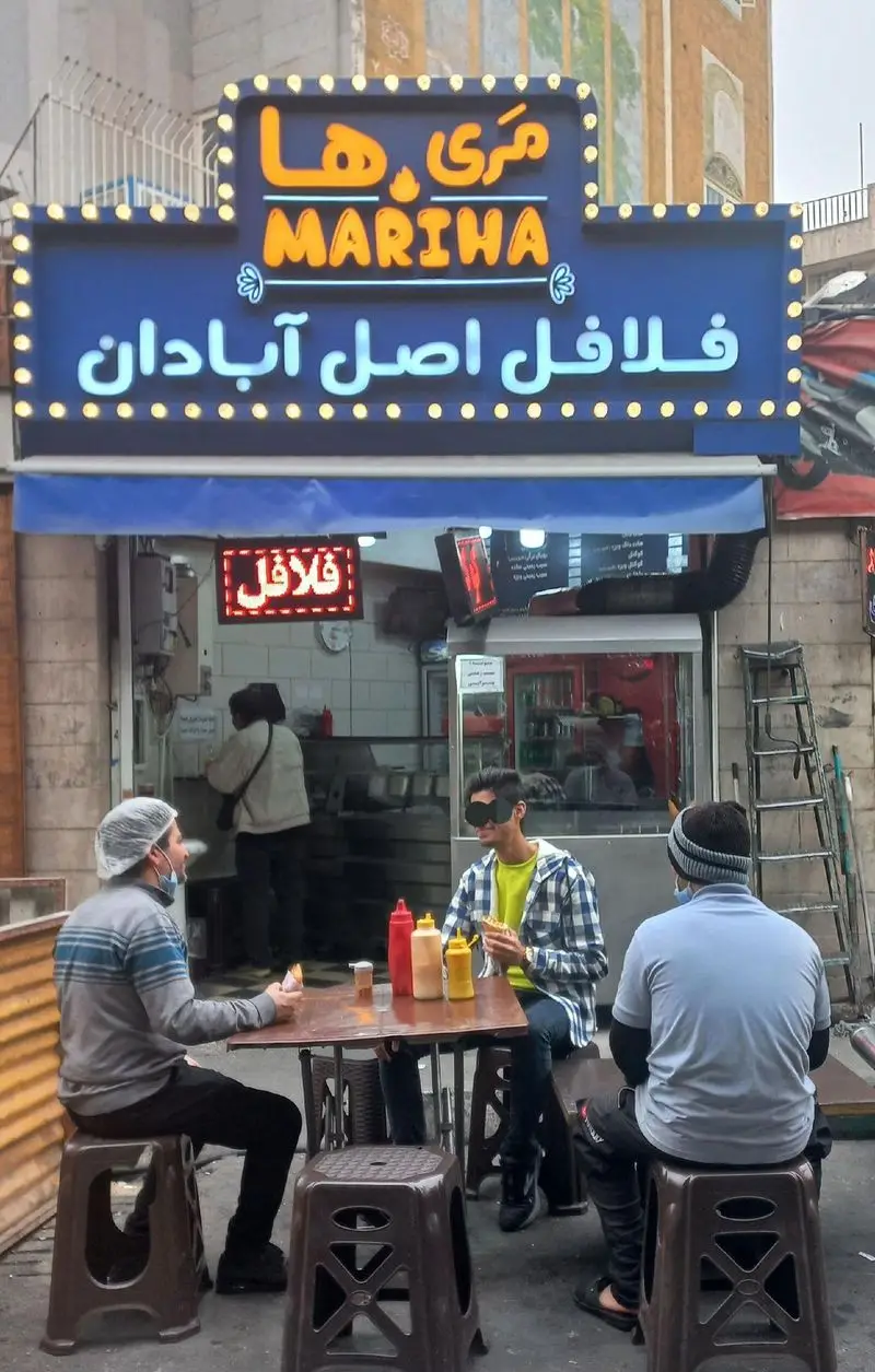تصویری از کسب و کار فست فود مَری ها با تابلوی درخشان "فلافل اصل آبادان" و مشتریانی که در فضای باز مشغول صرف غذا هستند.