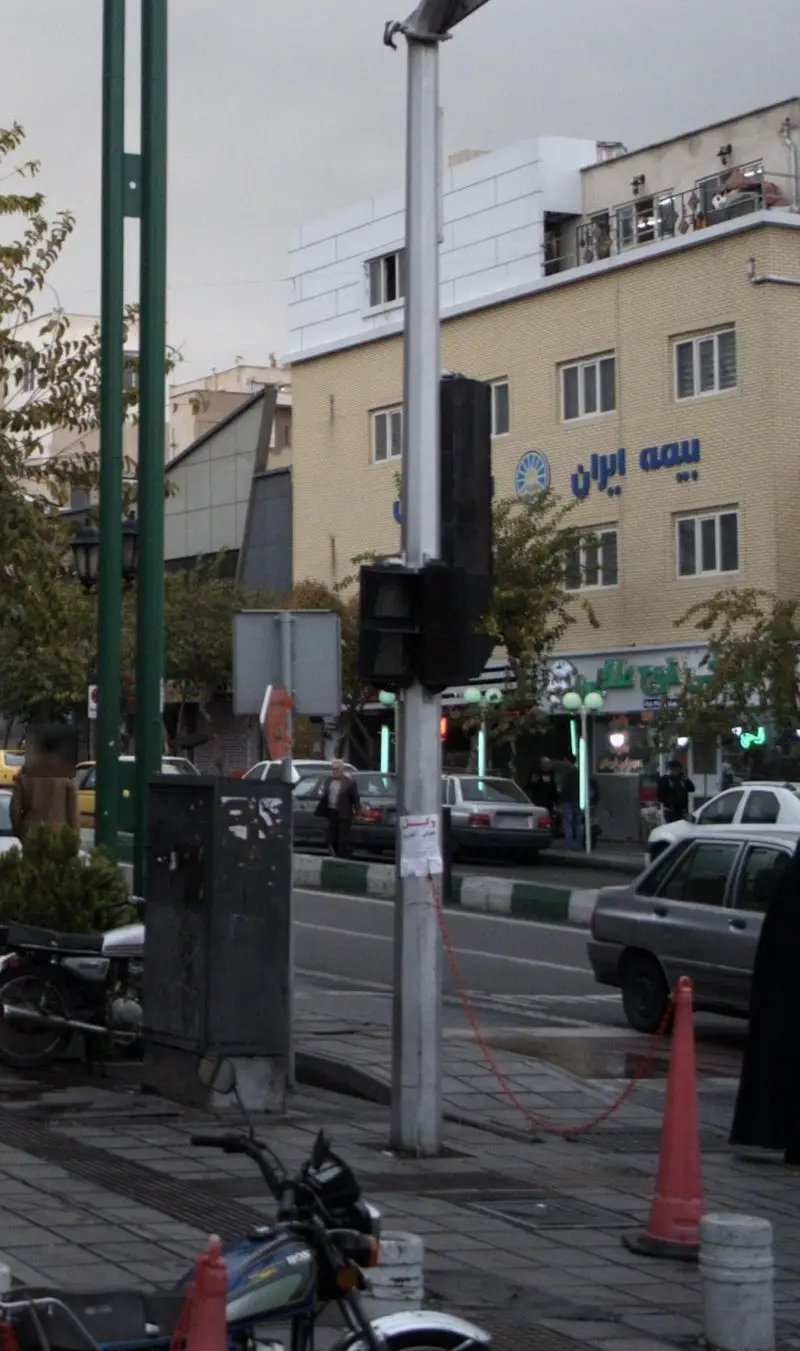 نمای خیابان پرتردد شهری با ساختمان بیمه ایران، خودروها و عابران پیاده، به همراه طباخی قوچ طلایی در میان کسب و کارهای محلی.