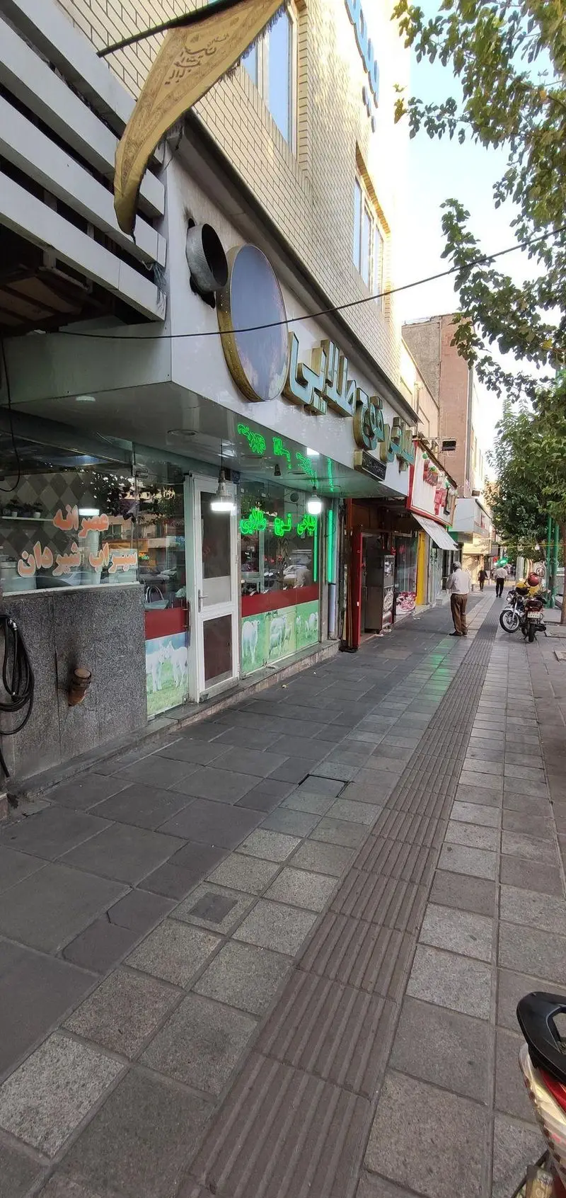 نمای بیرونی طباخی قوچ طلایی، مغازه کله پاچه فروشی سنتی در خیابانی شلوغ، با تابلوهای نئونی سبز، فرش تزئینی آویزان و عابران.