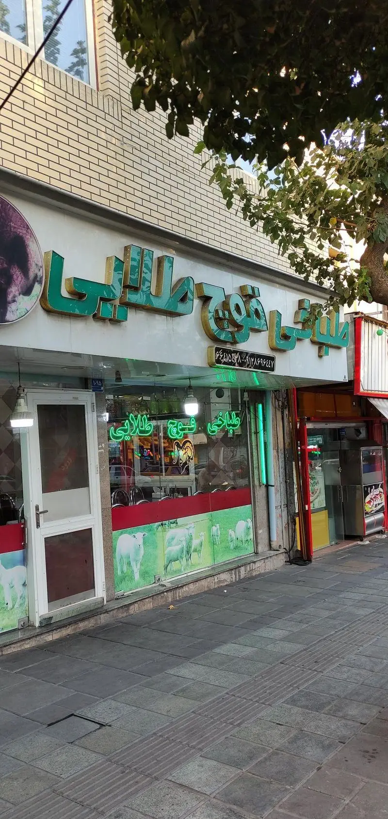 نمای بیرونی طباخی قوچ طلایی با تابلوی سبز رنگ برجسته، ویترین مزین به تصاویر گوسفندان و پیاده‌رو مقابل مغازه.