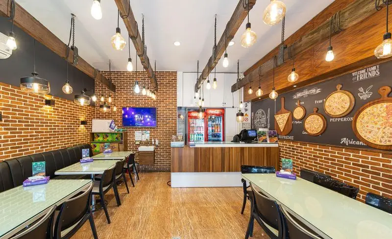 فضای داخلی مدرن و دلنشین فست فود سیب ۳۶۰ استاد معین با دیوارهای آجری، میز، صندلی، نورپردازی گرم و تابلو پیتزا.