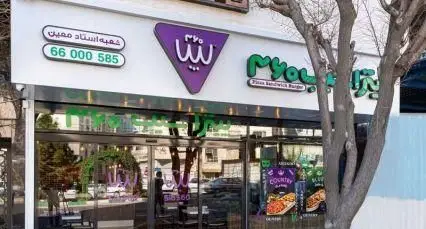 نمای بیرونی فست فود سیب ۳۶۰ شعبه استاد معین با تابلوهای روشن سبز و بنفش، ورودی شیشه‌ای، و خدمات پیتزا، ساندویچ و برگر.