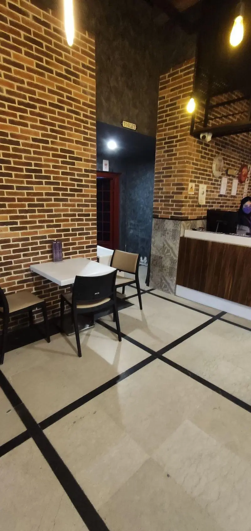 فضای داخلی فست فود سیب ۳۶۰ استاد معین با دکوراسیون آجری، میز و صندلی‌های مدرن، کانتر پذیرش و ورودی سرویس بهداشتی.