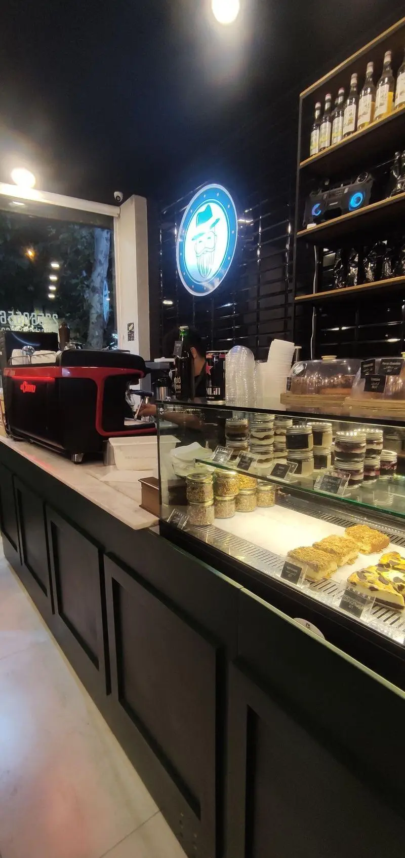 تصویر داخلی کافه قهوه سربن: دستگاه اسپرسو مشکی-قرمز، ویترین دسرهای شیک و شیرینی‌های تازه، لوگوی برند نورانی و قفسه بطری‌های متنوع.