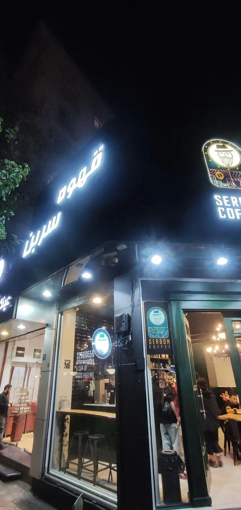 نمای شبانه از کافه قهوه سربن (Serbon Coffee) با تابلوهای درخشان، شماره تماس 22560556، لوگوی مرد ریشو و شعار "A Cup of Good Feeling".