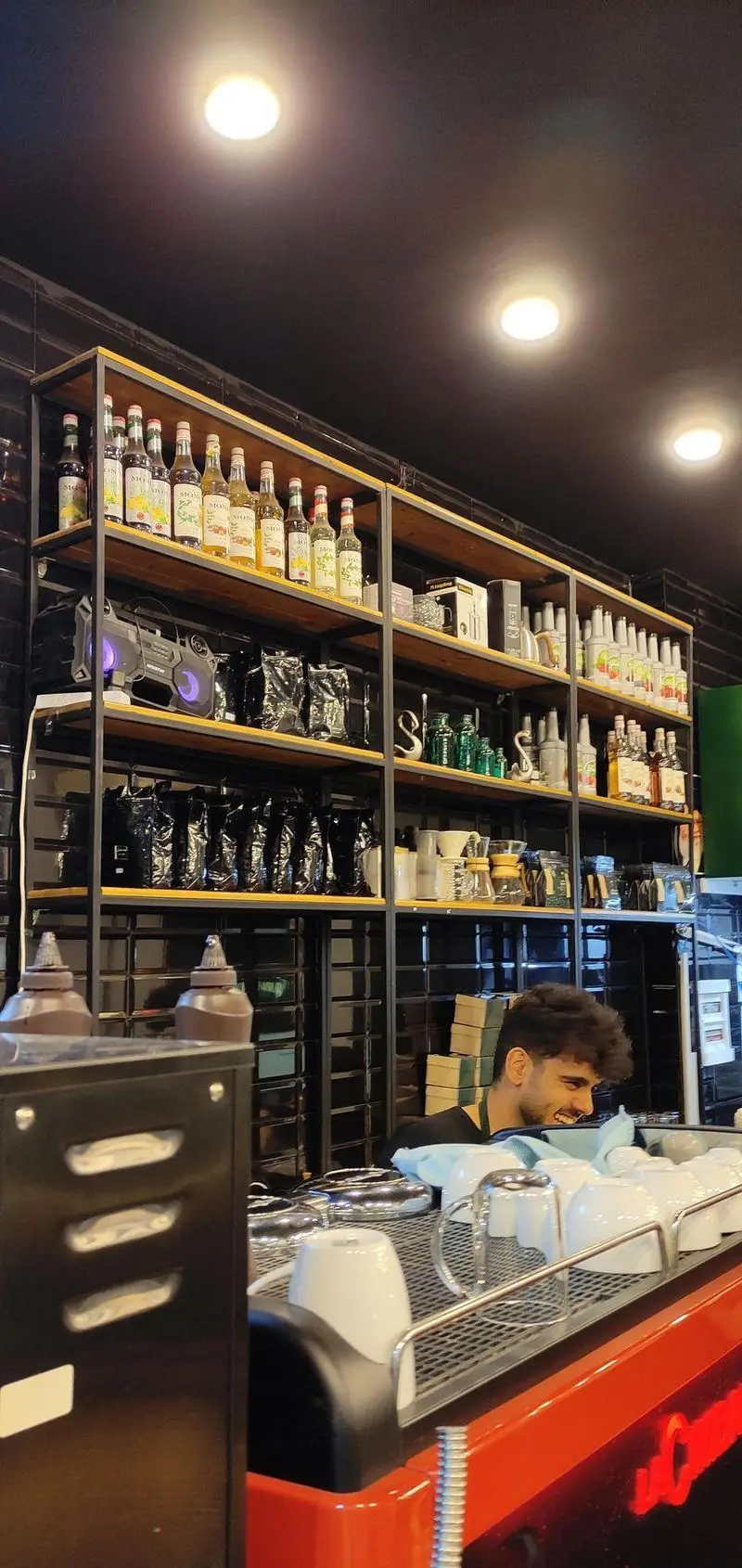 کافی شاپ قهوه سربن: قفسه‌های منظم با شربت‌های Monin، بسته‌های قهوه و لوازم باریستا. باریستای خوش‌رو در حال کار