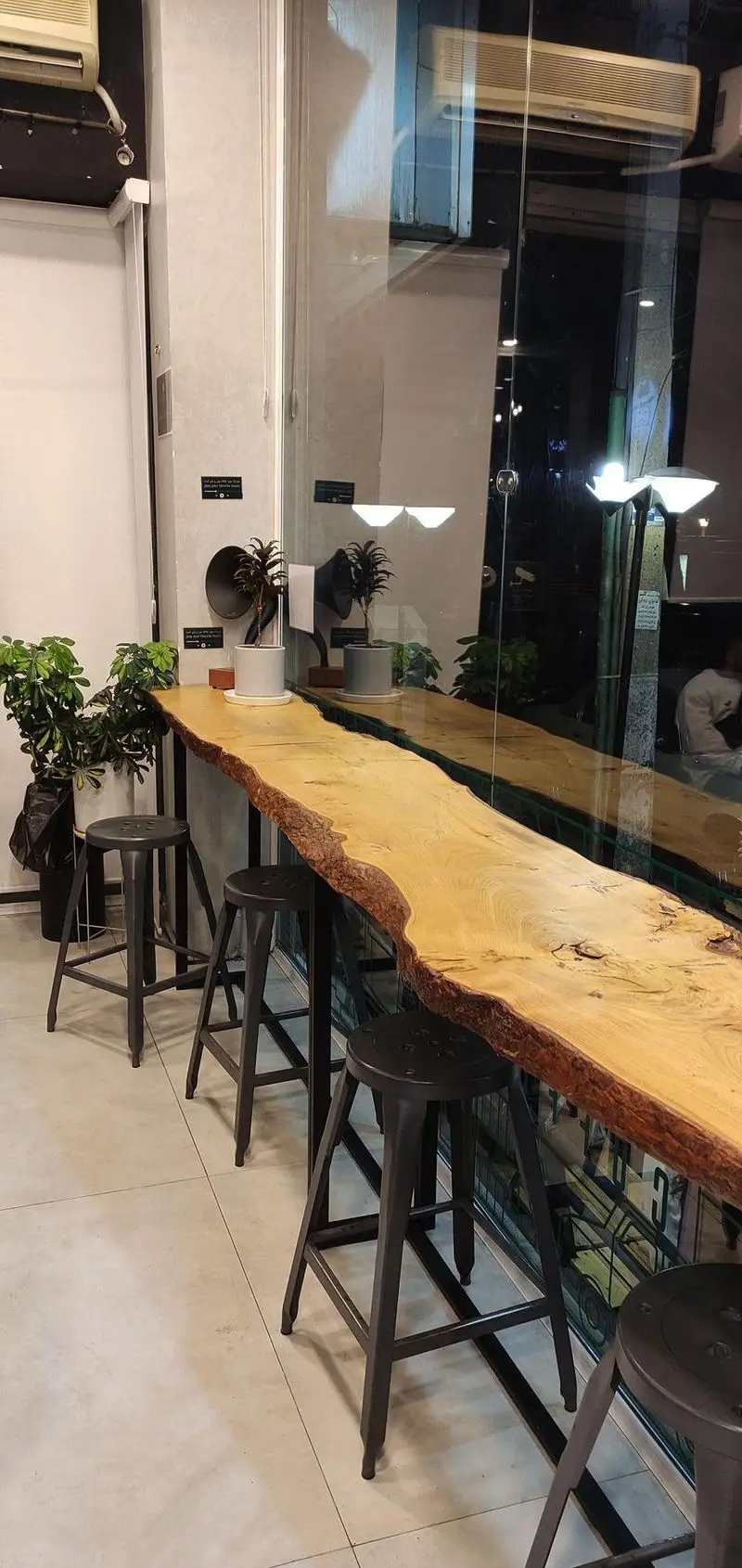 فضای داخلی مدرن و زیبای کافه قهوه سربن با میز چوبی روستیک، صندلی‌های کانتر شیک و گیاهان سبز. مکانی دنج برای نوشیدن قهوه.