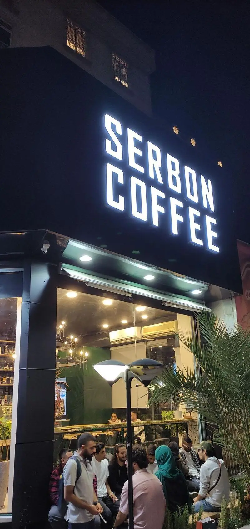 کافه قهوه سربن در شب، نمایی جذاب از نمای بیرونی با تابلوی نئون روشن و مشتریانی که در فضای دنج داخلی و خارجی دور هم جمع شده‌اند.