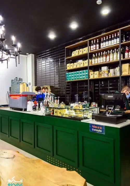 نمای داخلی کافی شاپ قهوه سربن با پیشخوان سبز رنگ، باریستاهای حرفه‌ای، دستگاه اسپرسو، قفسه‌هایی از انواع قهوه، شربت و شیرینی‌های تازه، آماده پذیرایی.
