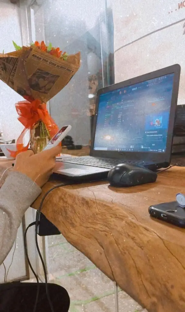 کافه‌ایندرای زیبا با میزی چوبی و لپ‌تاپ روشن، گوشی در دست و دسته‌گلی جذاب. محیطی مناسب کار و آرامش‌بخش برای مشتریان.