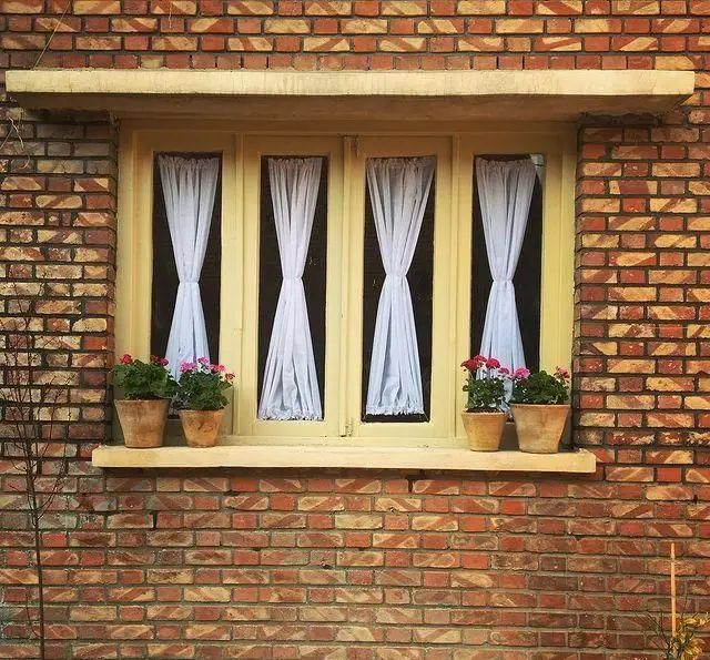 پنجره زیبای یک عمارت آجری با گلدان‌های گل رنگارنگ و پرده‌های سفید، نمادی از فضای دنج و دلنشین کافه عمارت نیلی.