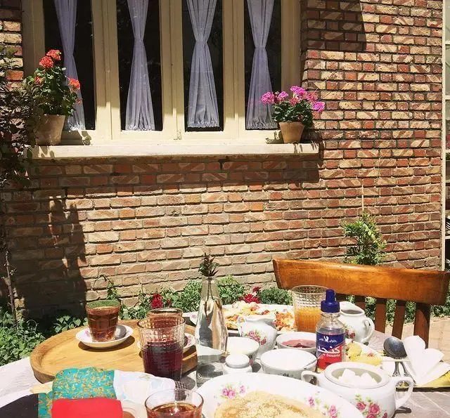 صبحانه دلنشین در فضای باز کافه عمارت نیلی، میز پر از غذا و نوشیدنی کنار دیوار آجری و پنجره های زیبا با گلدان گل.