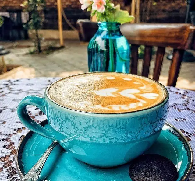 فنجان لاته آرت آبی فیروزه‌ای زیبا با طرح برگ، روی میز گیپور کافه عمارت نیلی. تجربه قهوه در فضای دلنشین و باز این کافه.