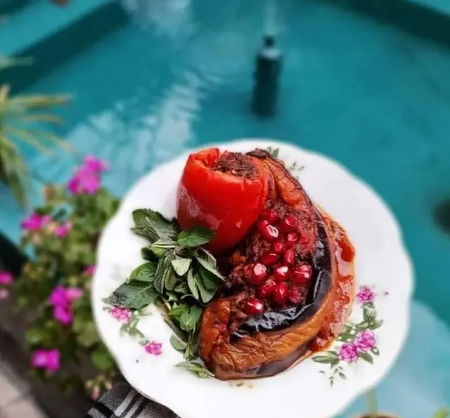 غذای سنتی دلمه بادمجان و فلفل شکم پر با تزئین انار و سبزیجات تازه، سرو شده در فضای دلنشین و سرسبز کافه عمارت نیلی.