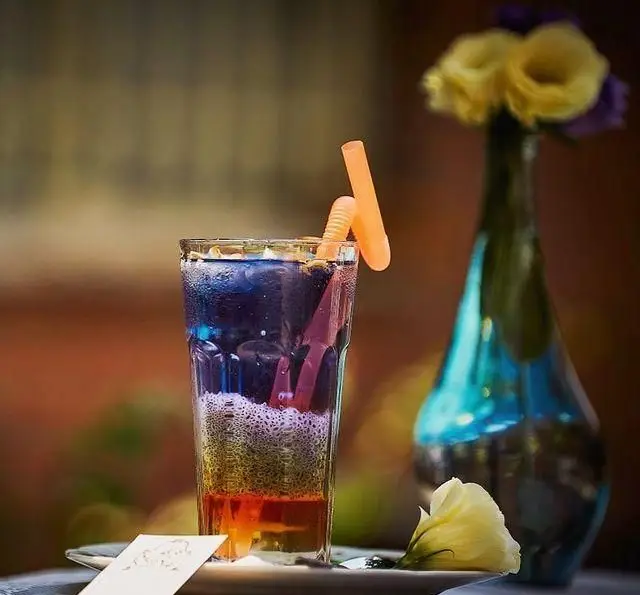 نوشیدنی خنک و لایه‌لایه کافه عمارت نیلی با رنگ‌های آبی، سبز و نارنجی، شامل دانه‌های خوراکی، کف لطیف و نی برای روزهای گرم.